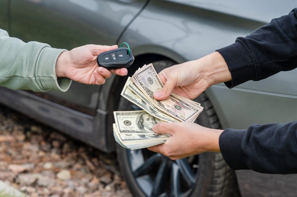 Two people exchanging money and car keys, with one person handing over a stack of cash and the other holding a car key, standing next to a vehicle